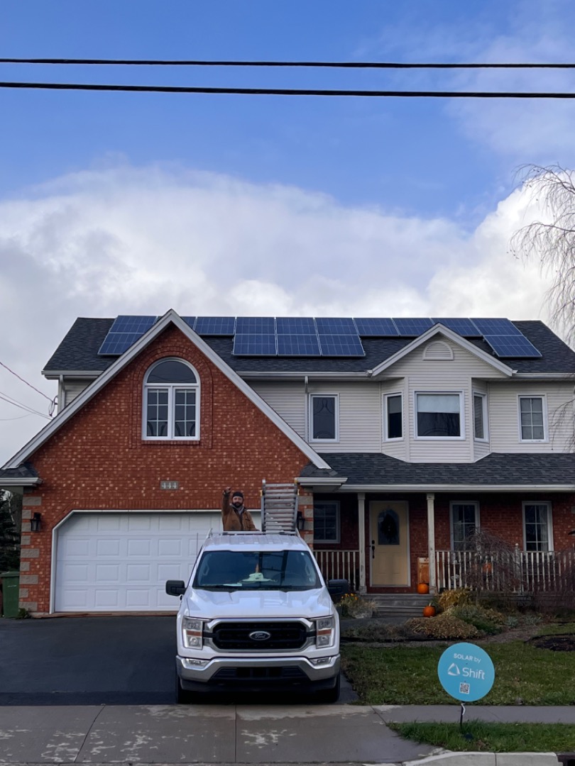 Front view of a home with solar panel installed on the roof. There is a Shift truck in the driveway, with a solar installer in the back giving a thumbs-up, and a Shift lawn sign in the grass.