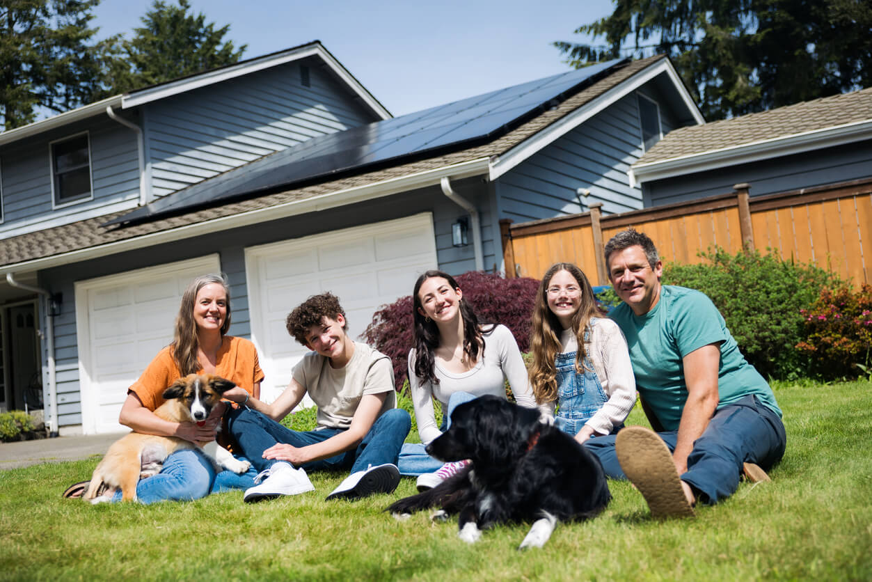 Family of 5 with their dog, sitting in their front lawn. Their home and solar panels are visible in the background.