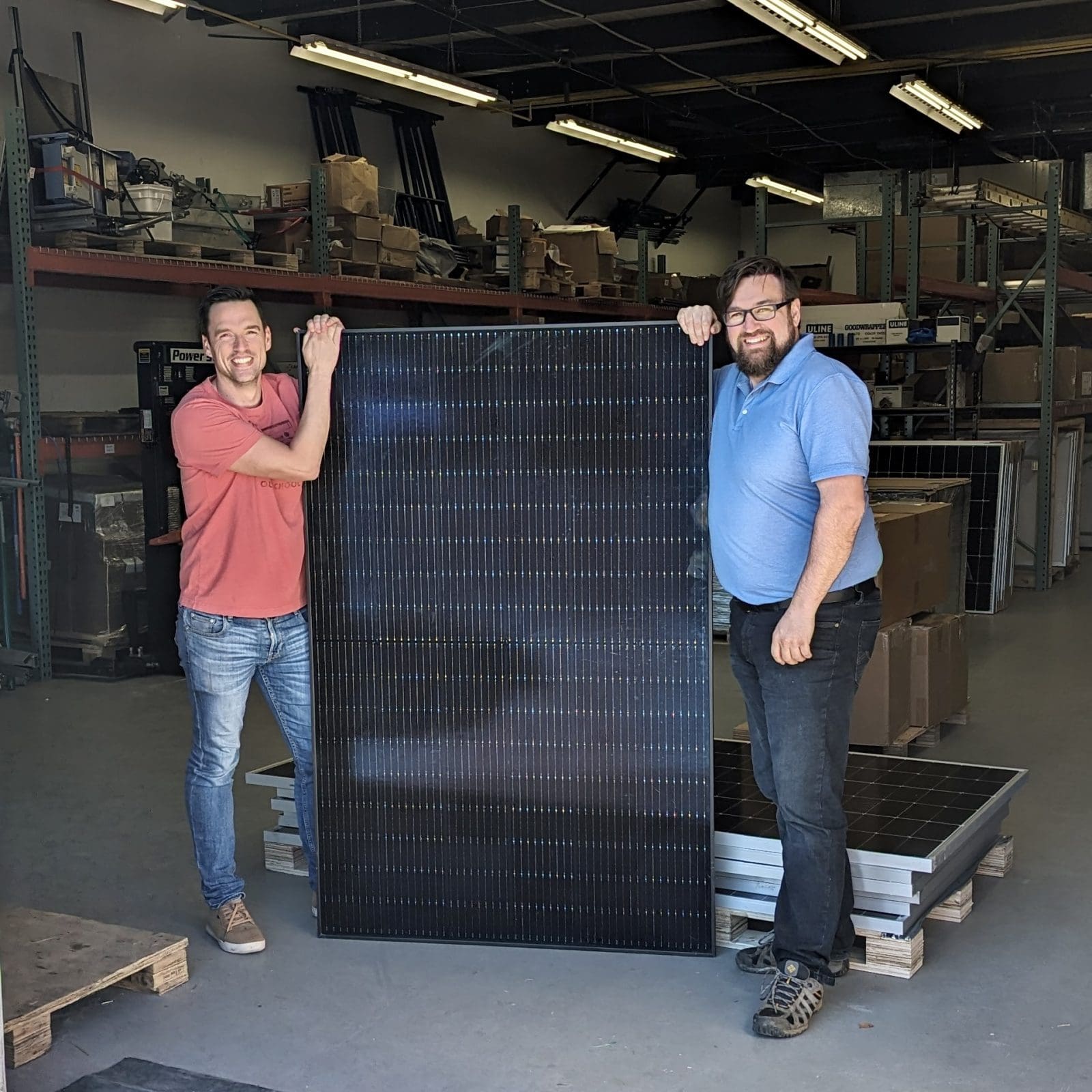 2 men standing inside a warehouse, on either side of a solar panel they are posing with.