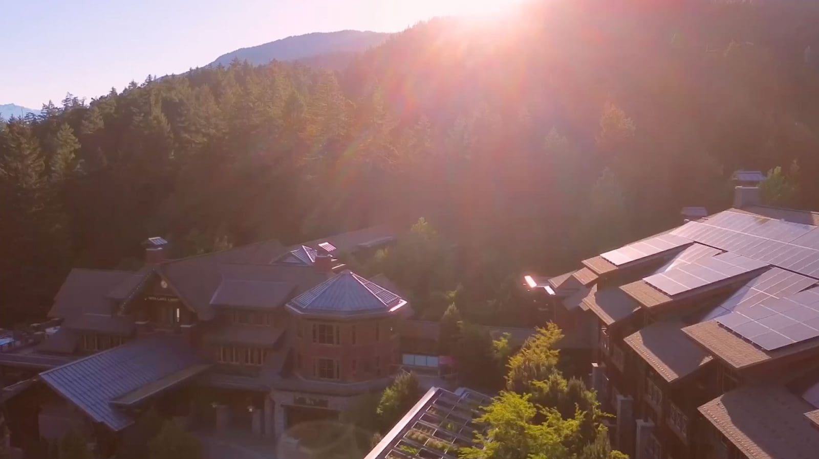 An aerial view of Nita Lake Lodge. There is a large solar panel installation on the roof. In the back ground, the lodge is surrounded by trees and the sun is setting behind a mountain.
